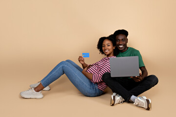 Cheerful young black man and lady with big belly sit on floor, enjoy pregnancy with laptop, show credit card