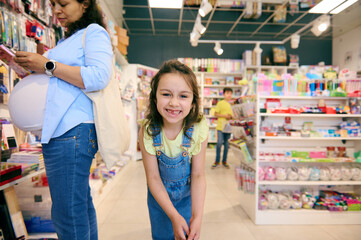 Adorable mischievous little child girl smiling looking at camera, shopping with her mother in the school stationery shop