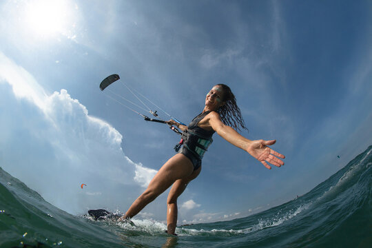 Kitesurfing girl in black sexy swimsuit with kite in sky on board in blue sea riding waves with water splash. Recreational activity, water sports, action, hobby and fun in summer time. 