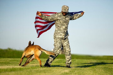 Soldier in military uniform holding USA flag and celebrating freedom and patriotism with his dog.