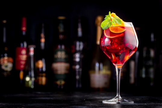 Red sangria summer alcoholic drink with red spanish wine, peach, plum, orange, lime and ice. Black bar counter background, steel bar tools and bottles