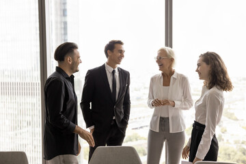 Happy younger and older business colleagues in formal clothes standing together at large office window, talking, laughing, discussing successful project, creative ideas, relaxing on work break