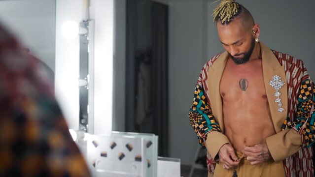 Attractive stylish black gay man wear ethnic jacket look at mirror view from behind. Lifestyle lgbtq pride concept. Feminine man with beard and makeup on face straightens clothes in front of mirror