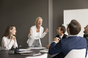 Cheerful senior business leader woman and team of excited younger employees meeting at table, brainstorming on project, discussing creative ideas, laughing out loud, having fun