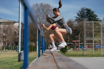 Fit sportsgirls doing jumps on the bench while training outdoors in the park, on a sunny day
