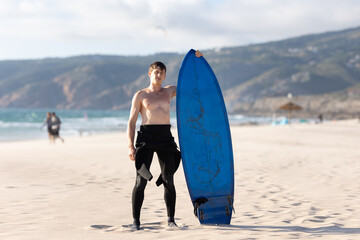 Fototapeta na wymiar A man with naked torso standing on the beach holding a blue surfboard