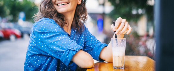 Caucasian younfg woman drinking iced coffee on the outdoor terrace of a cafe on a hot summer day.