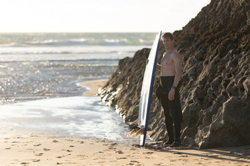 A smiling man with a naked torso standing by a rock holding a surfboard