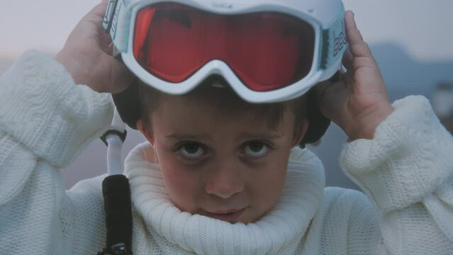 Cute Caucasian boy puts on a protective helmet for skiing or snowboarding. Portrait of a pretty child in the mountains, wearing glasses or a red mask. Winter cozy concept. Son's big blue eyes