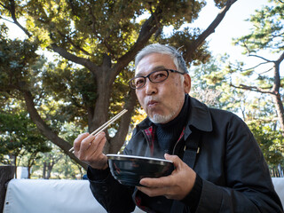 An older asian man eating noodles and soup with chopsticks - 629243842