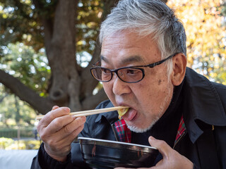 An older asian man eating noodles and soup with chopsticks - 629244281