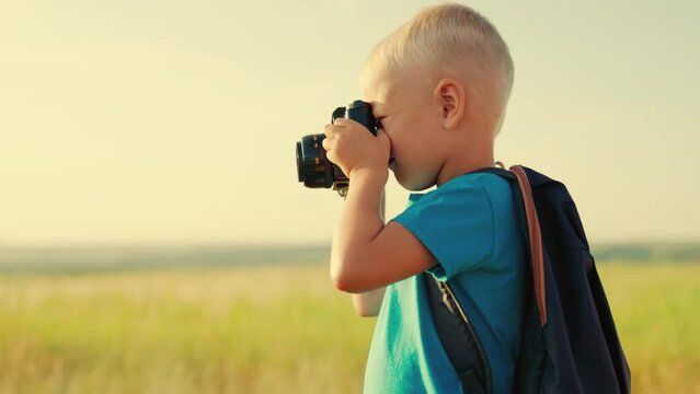Child with camera take pictures of nature on summer day. Boy with backpack on back looks into camera lens, takes beautiful photo greenfield. Kids with video camera in fresh air films about nature