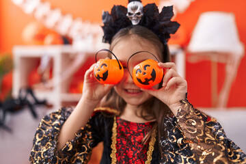 Adorable hispanic girl wearing halloween costume covering eyes with small pumpkin baskets at home