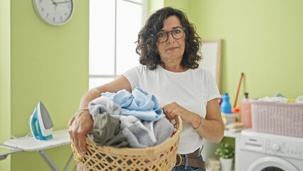 Middle age hispanic woman holding basket with clothes tired at laundry room