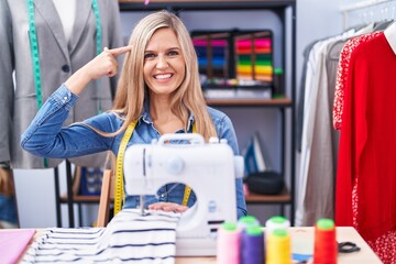 Blonde woman dressmaker designer using sew machine smiling pointing to head with one finger, great idea or thought, good memory