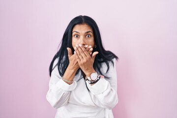 Mature hispanic woman standing over pink background shocked covering mouth with hands for mistake....