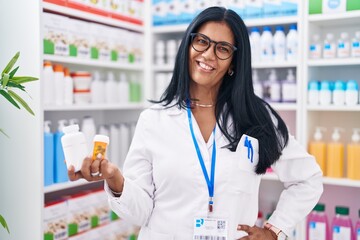 Middle age hispanic woman pharmacist smiling confident holding pills bottles at pharmacy