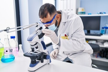Young hispanic man scientist wearing medical mask using microscope at laboratory