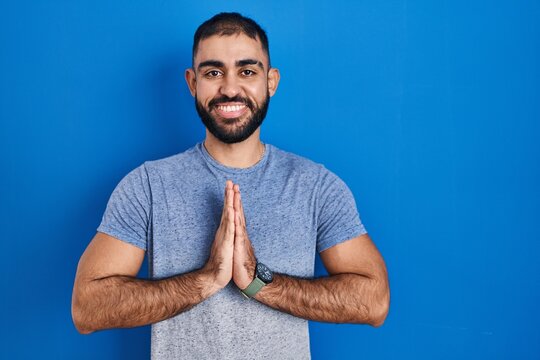Middle east man with beard standing over blue background praying with hands together asking for forgiveness smiling confident.