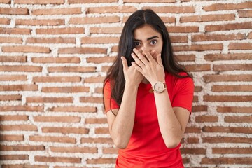 Young teenager girl standing over bricks wall shocked covering mouth with hands for mistake. secret concept.