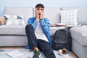 Young hispanic man sitting on the floor studying for university afraid and shocked, surprise and...