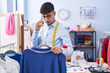 Young hispanic man tailor talking on smartphone holding t shirt at tailor shop