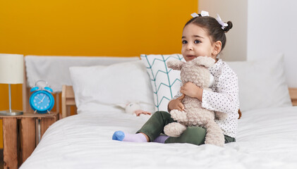 Fototapeta premium Adorable hispanic girl hugging teddy bear sitting on bed at bedroom