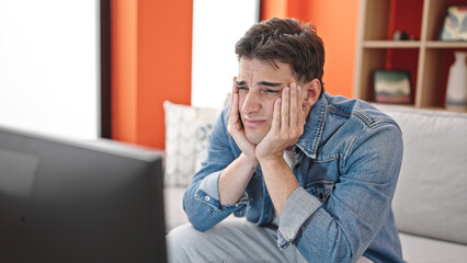 Young hispanic man watching tv sitting on sofa with boring expression at home