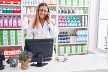 Young beautiful hispanic woman pharmacist talking on smartphone using computer at pharmacy