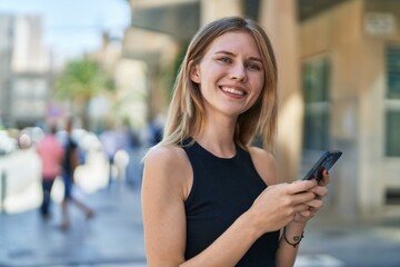 Young blonde woman using smartphone smiling at street