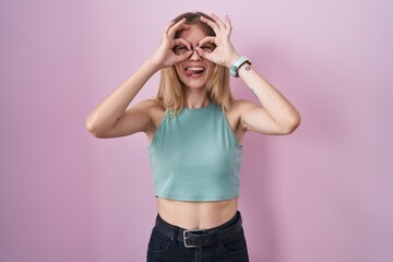 Obraz na płótnie Canvas Blonde caucasian woman standing over pink background doing ok gesture like binoculars sticking tongue out, eyes looking through fingers. crazy expression.