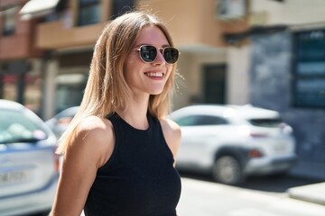 Young blonde woman wearing sunglasses looking to the side smiling at street