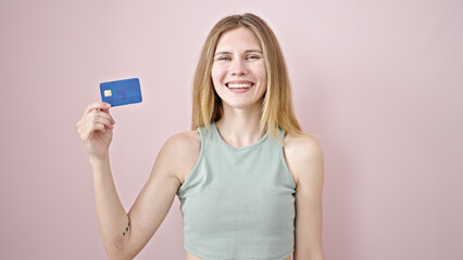 Naklejka premium Young blonde woman smiling confident holding credit card over isolated pink background