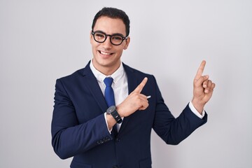 Young hispanic man wearing suit and tie smiling and looking at the camera pointing with two hands and fingers to the side.