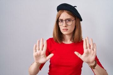 Obraz na płótnie Canvas Young redhead woman standing wearing glasses and beret moving away hands palms showing refusal and denial with afraid and disgusting expression. stop and forbidden.