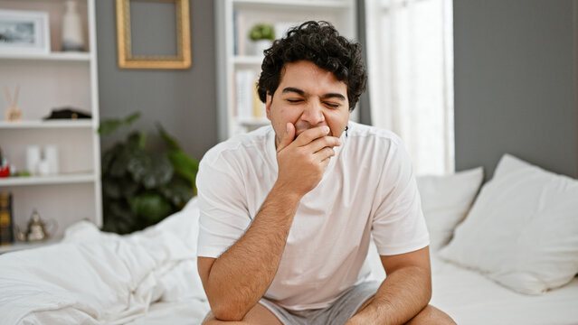 Young latin man sitting on bed yawning at bedroom
