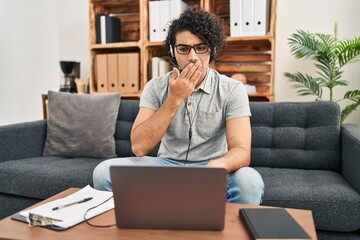 Hispanic man with curly hair doing online session at consultation office covering mouth with hand, shocked and afraid for mistake. surprised expression