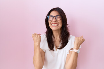 Middle age hispanic woman wearing casual white t shirt and glasses very happy and excited doing winner gesture with arms raised, smiling and screaming for success. celebration concept.