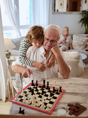 Loving grandson hugging grandfather and playing chess at home