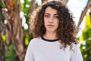 Hispanic woman with curly hair standing outdoors thinking attitude and sober expression looking...