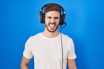 Hispanic man with beard listening to music wearing headphones sticking tongue out happy with funny...
