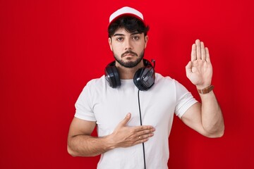 Hispanic man with beard wearing gamer hat and headphones swearing with hand on chest and open palm,...
