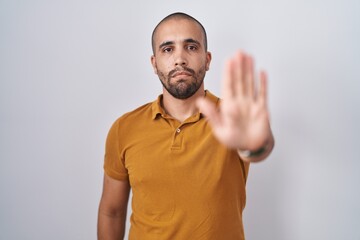 Hispanic man with beard standing over white background doing stop sing with palm of the hand....