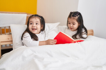 Adorable twin girls reading book sitting on bed at bedroom