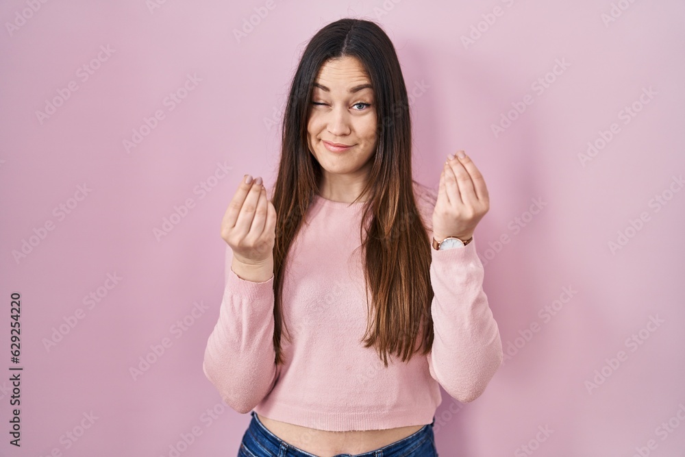 Sticker young brunette woman standing over pink background doing money gesture with hands, asking for salary