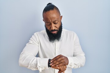 African american man standing over blue background checking the time on wrist watch, relaxed and...