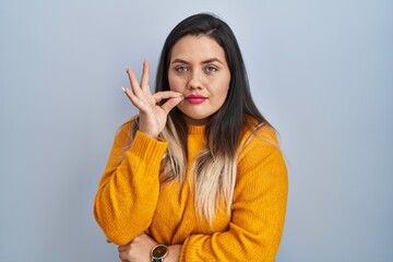 Young hispanic woman standing over isolated background mouth and lips shut as zip with fingers....