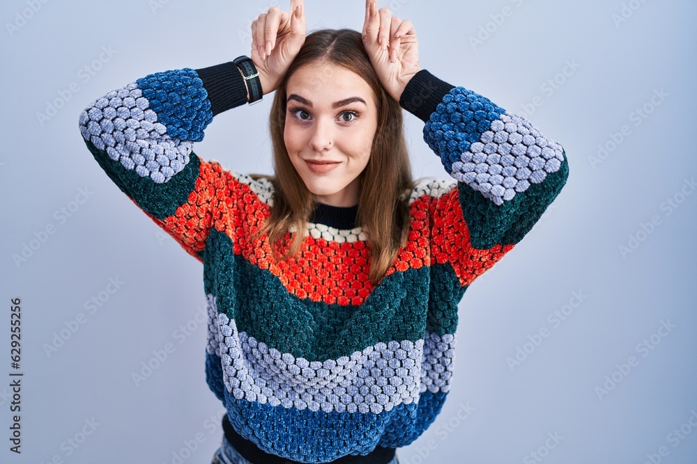 Sticker Young hispanic girl standing over blue background doing funny gesture with finger over head as bull horns