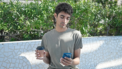 Young hispanic man using smartphone drinking coffee at park