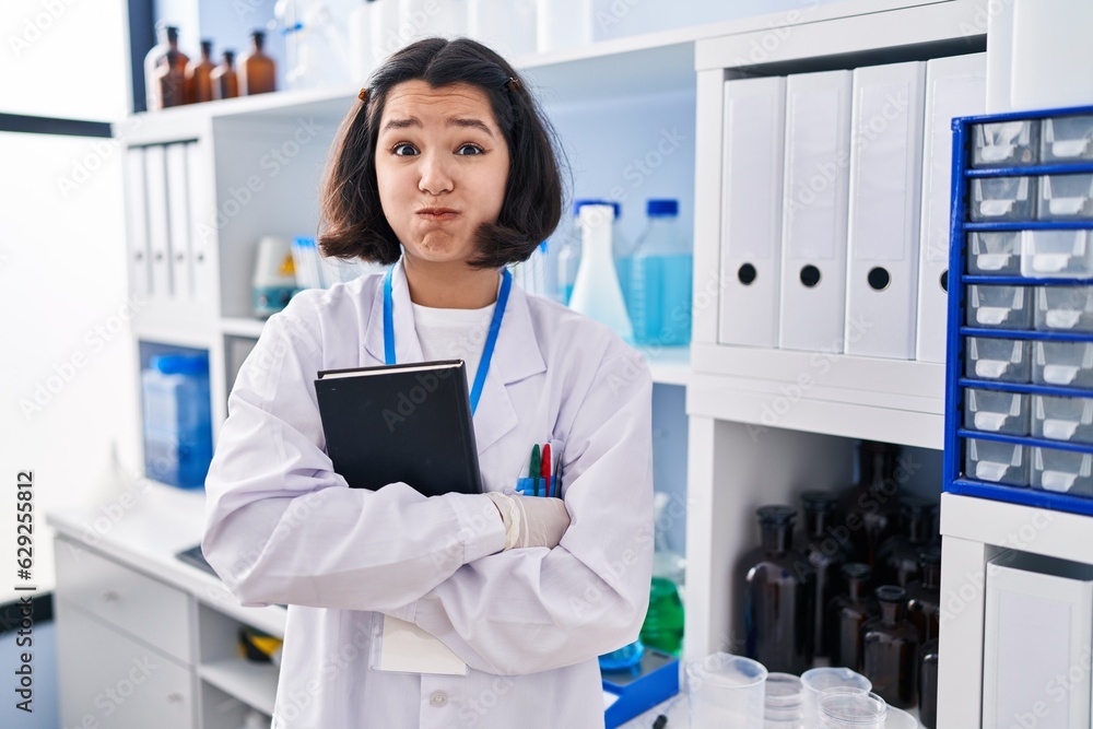 Sticker young hispanic woman working at scientist laboratory puffing cheeks with funny face. mouth inflated 